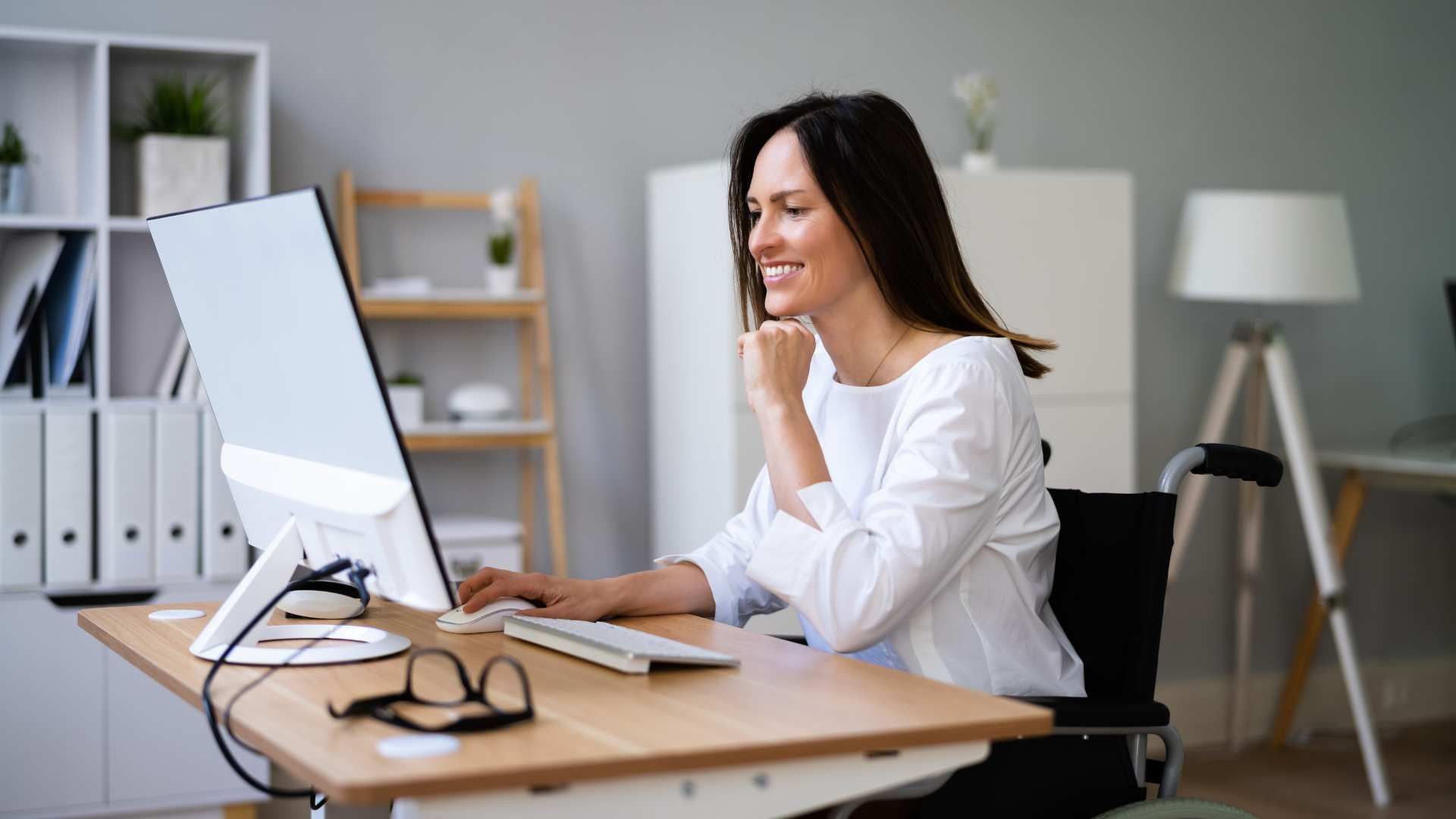 Woman at a desk using a desktop computer in a bright office, smiling thoughtfully.