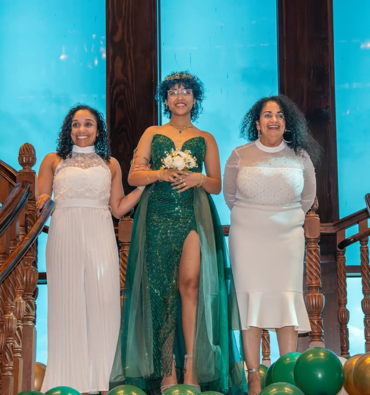 Three people stand on a staircase, with two wearing white dresses and one in a green sequined gown holding a bouquet.
