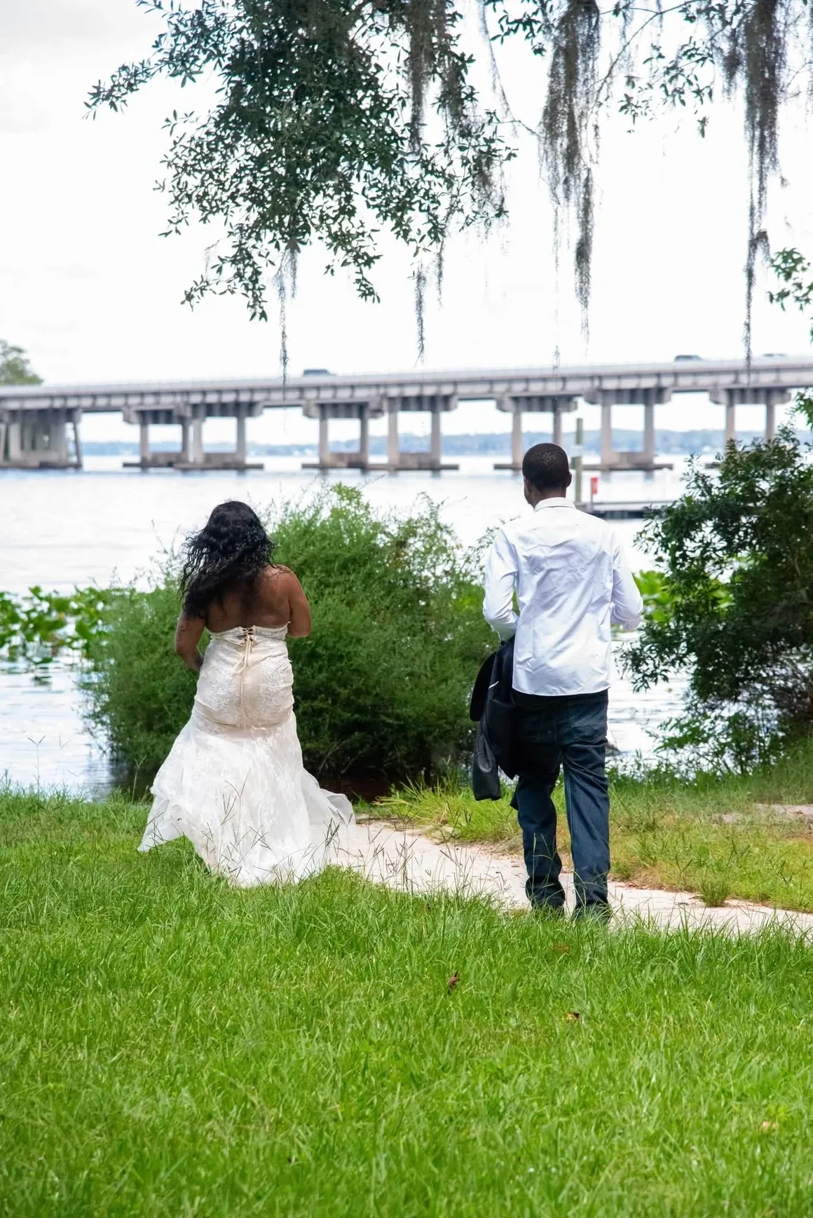 A couple in formal attire walks away from the camera along a grassy path toward a bridge over the water.