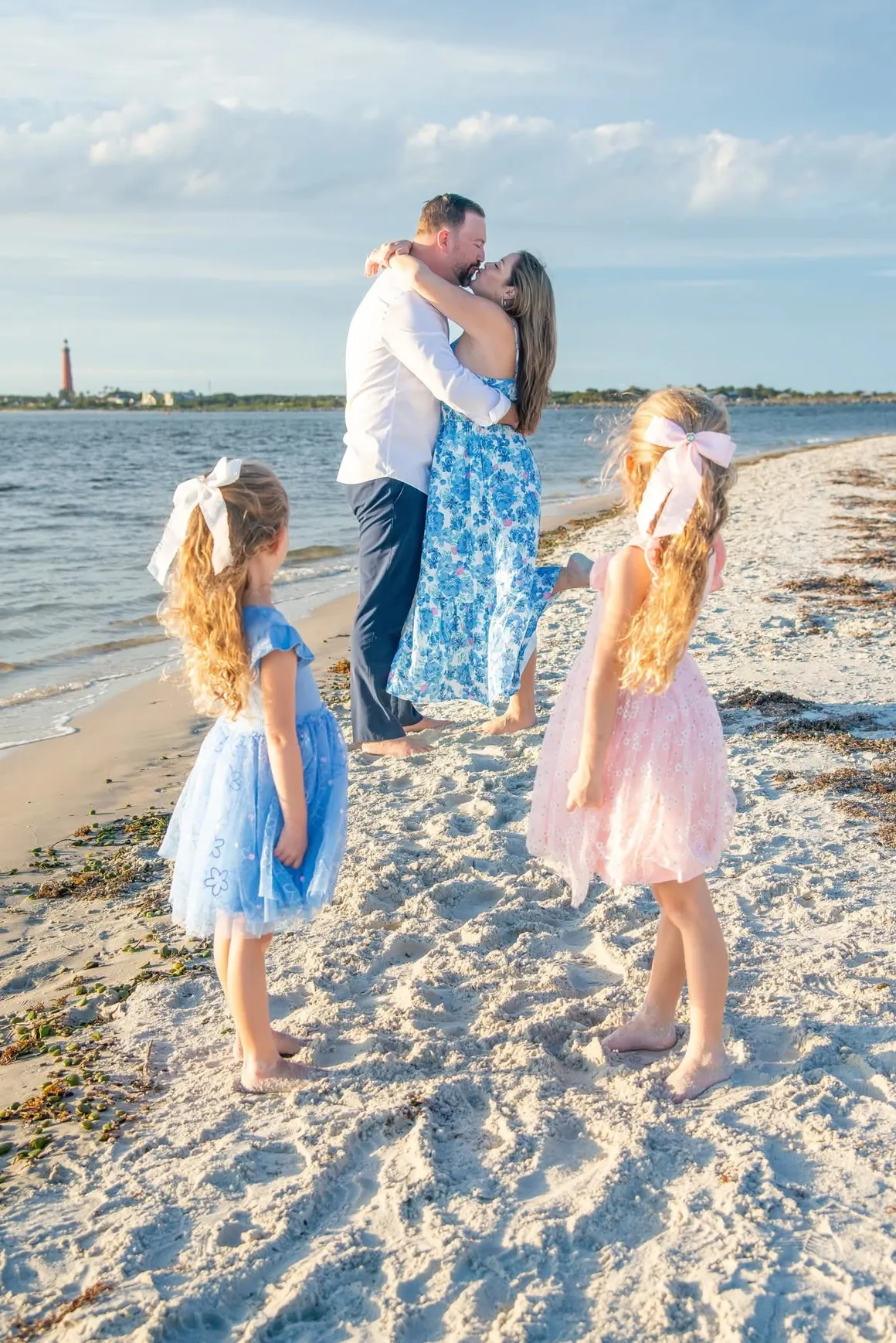 A family standing on a sandy beach, with two children in dresses looking toward a couple embracing by the water.