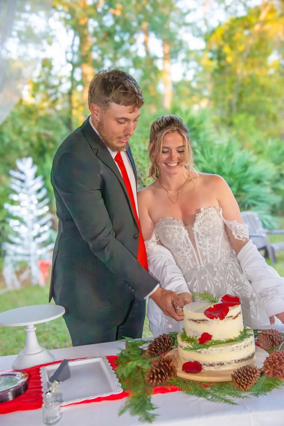 A couple in formal attire cuts a two-tiered wedding cake decorated with red flowers and pine cones outdoors.