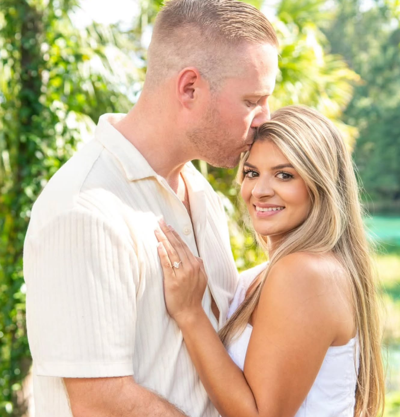 A person kisses another person's forehead outdoors in a sunlit, green setting. Both are dressed in white clothing.