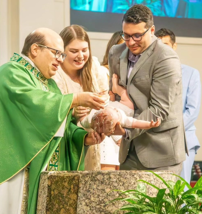 A priest baptizes an infant over a stone font as two parents look on with smiles in a church setting.