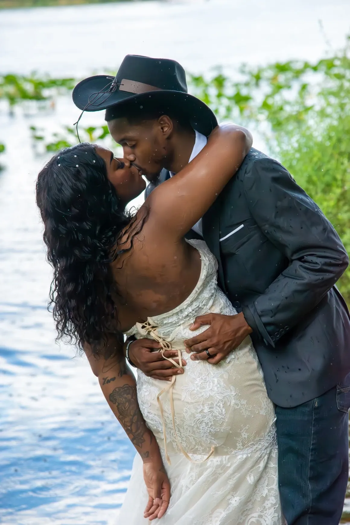 A couple in formal wedding attire, including a cowboy hat, kiss while embracing beside a lake.