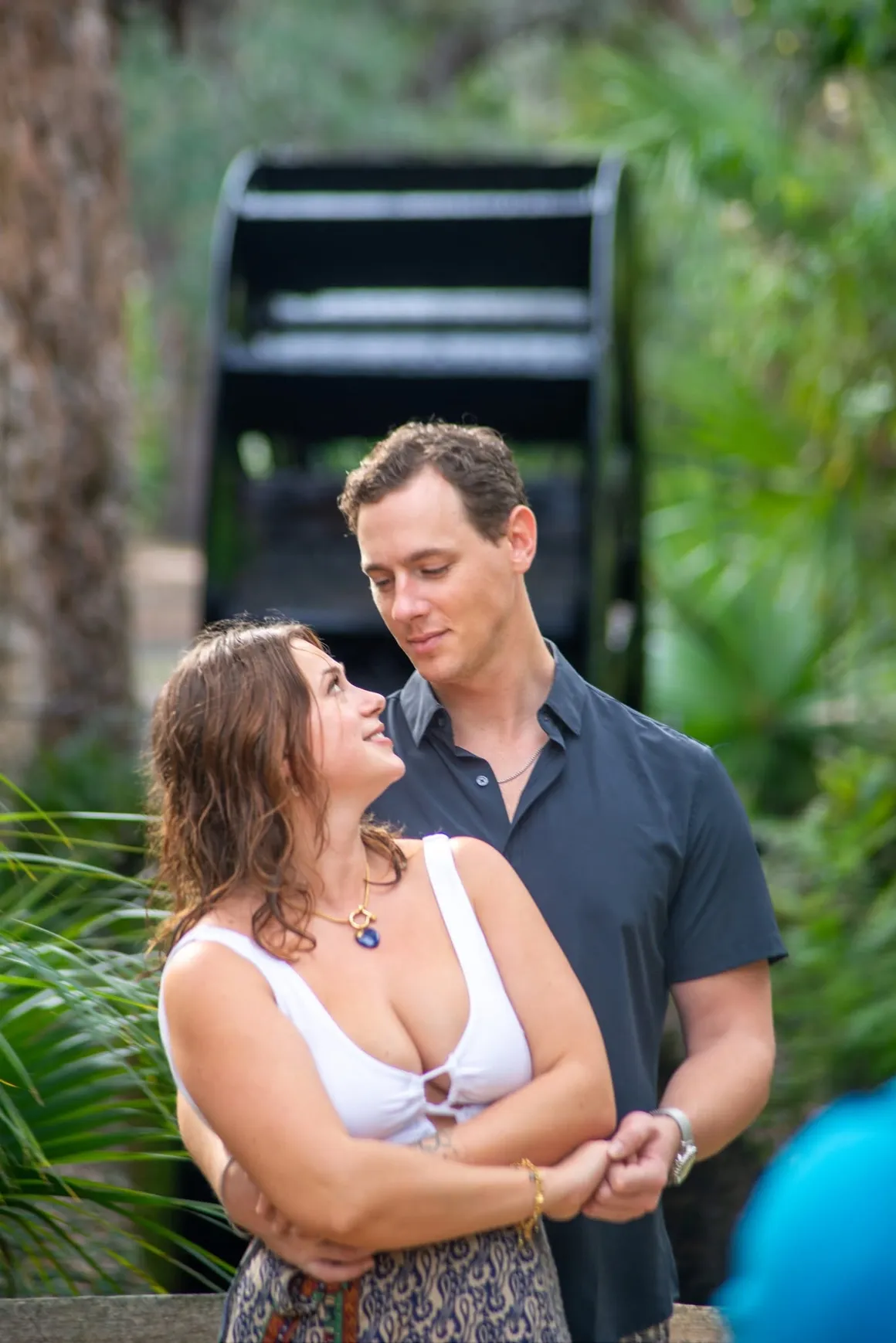 A couple stands together in a garden, gazing into each other’s eyes near a large, black wooden water wheel.