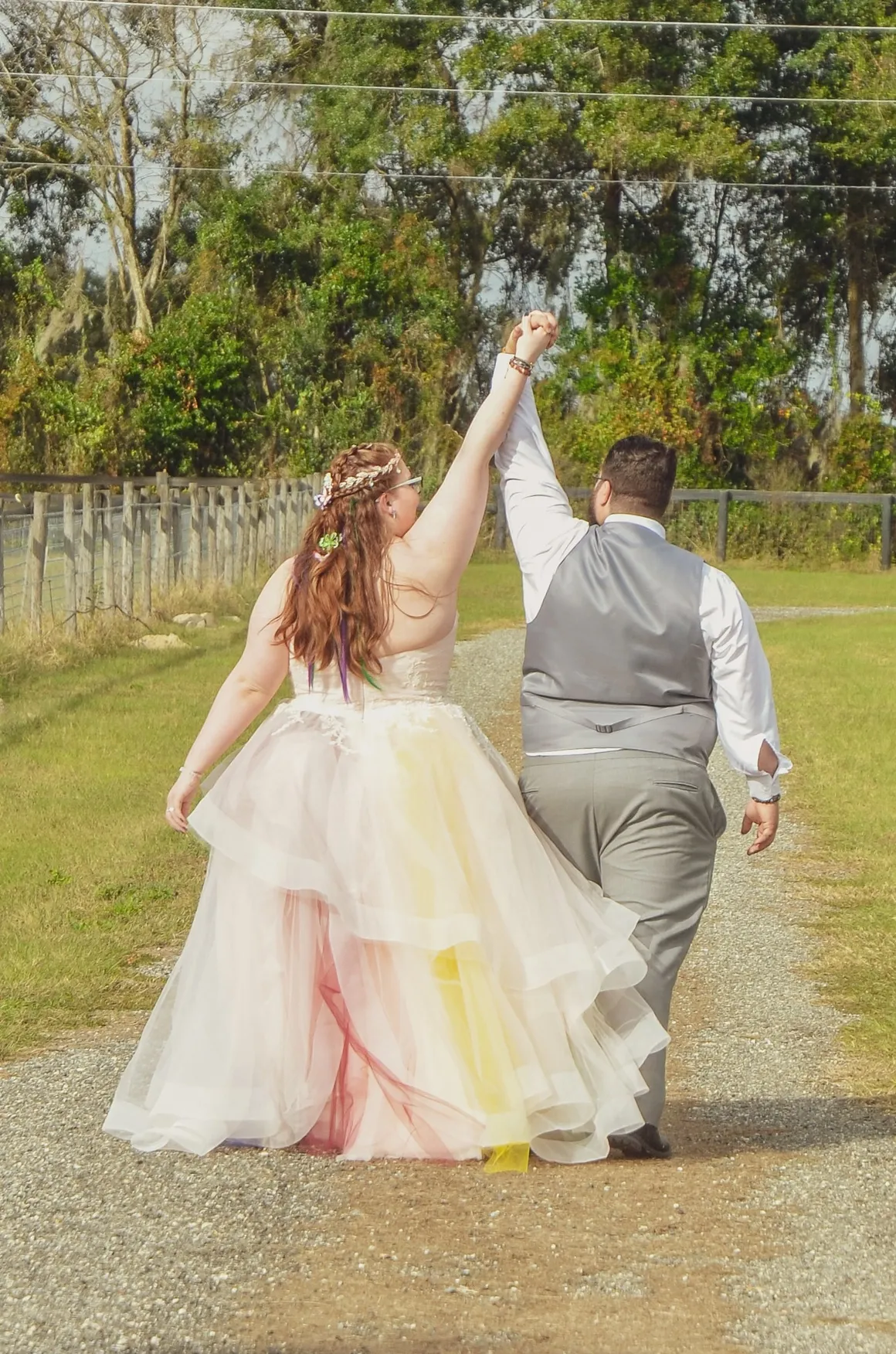A couple walks away on a gravel path, hands held high, wearing a multicolored tulle wedding gown and a gray suit vest.