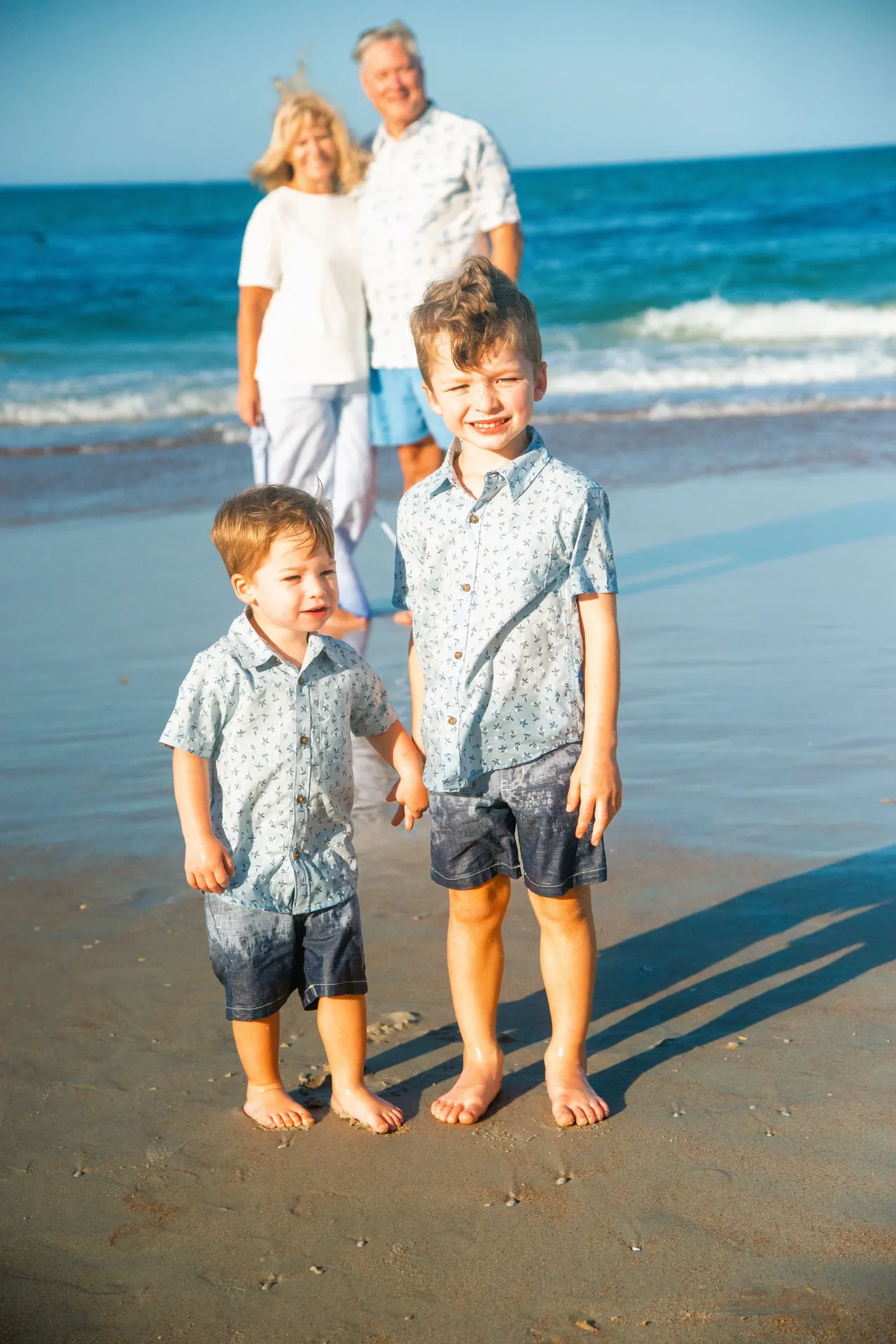 Two children in matching shirts stand on a beach in front of a pair of adults standing by the ocean.