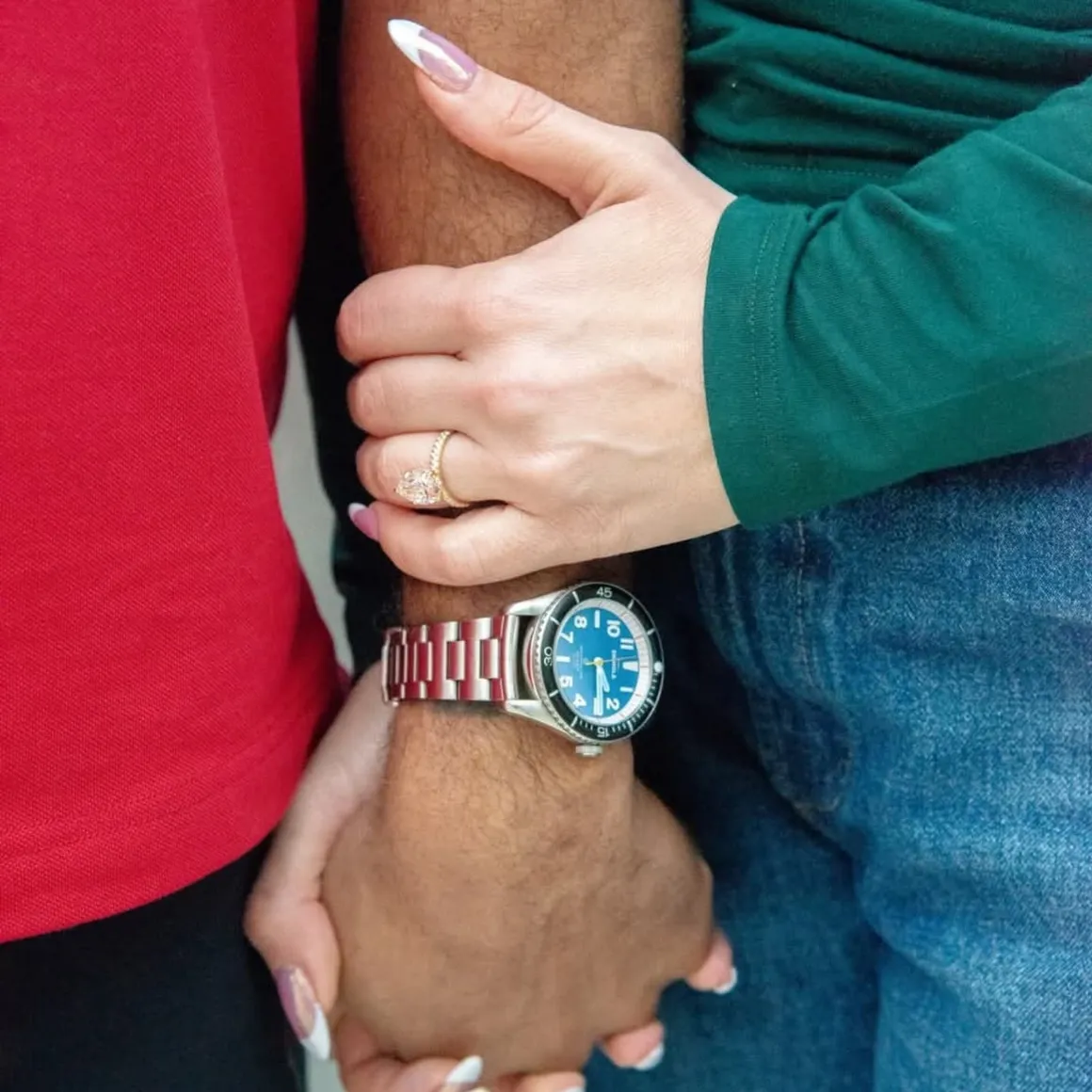 Two people holding hands, one wearing a watch with a blue face and metal band, and the other wearing a gold ring.
