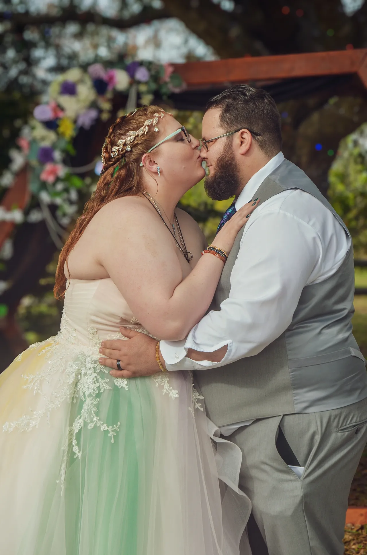 A couple shares a kiss at their outdoor wedding, the bride in a pastel-layered gown and the groom in a grey suit.