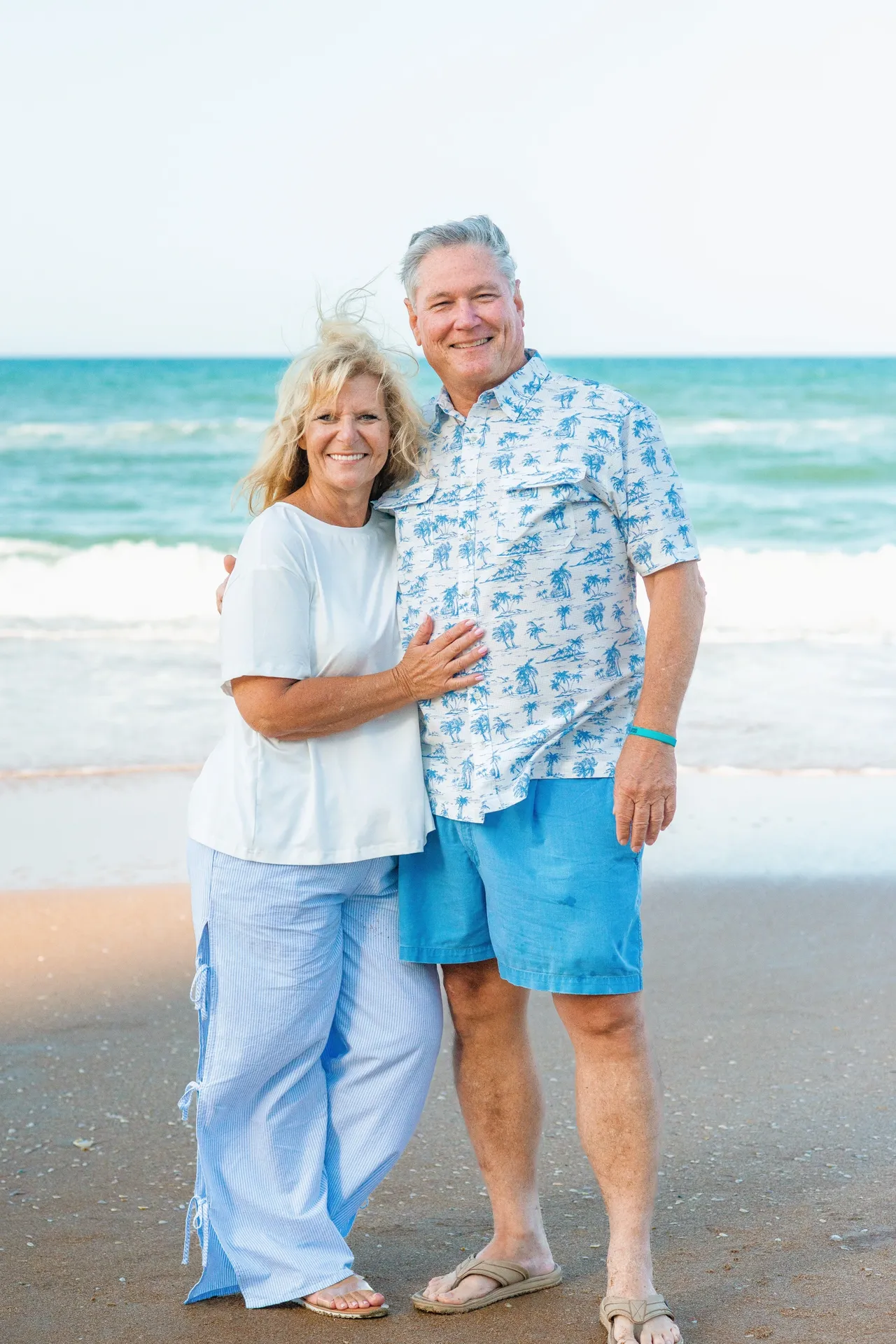 A smiling couple stands on a beach, posing together in light-colored beach attire with the ocean in the background.