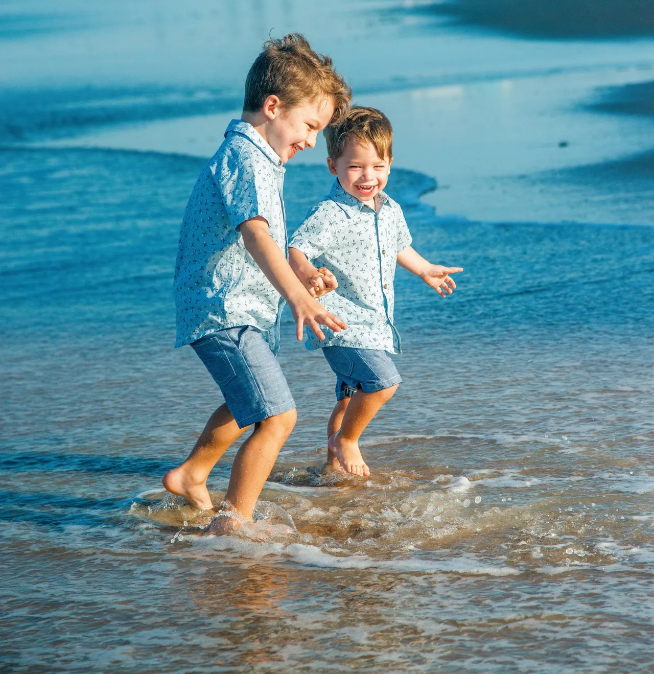 Two children in matching light blue patterned shirts and shorts run and splash in shallow ocean water on a sunny day.