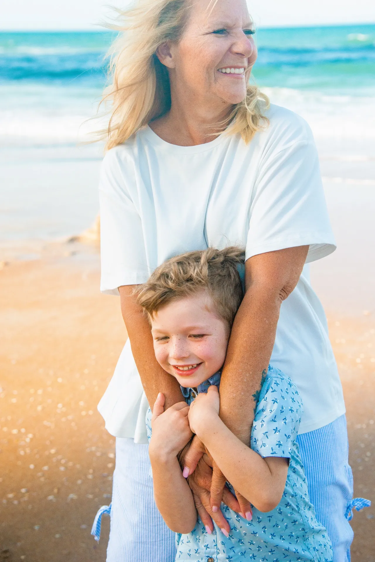 A person hugs a child from behind while standing on a sandy beach near the ocean.