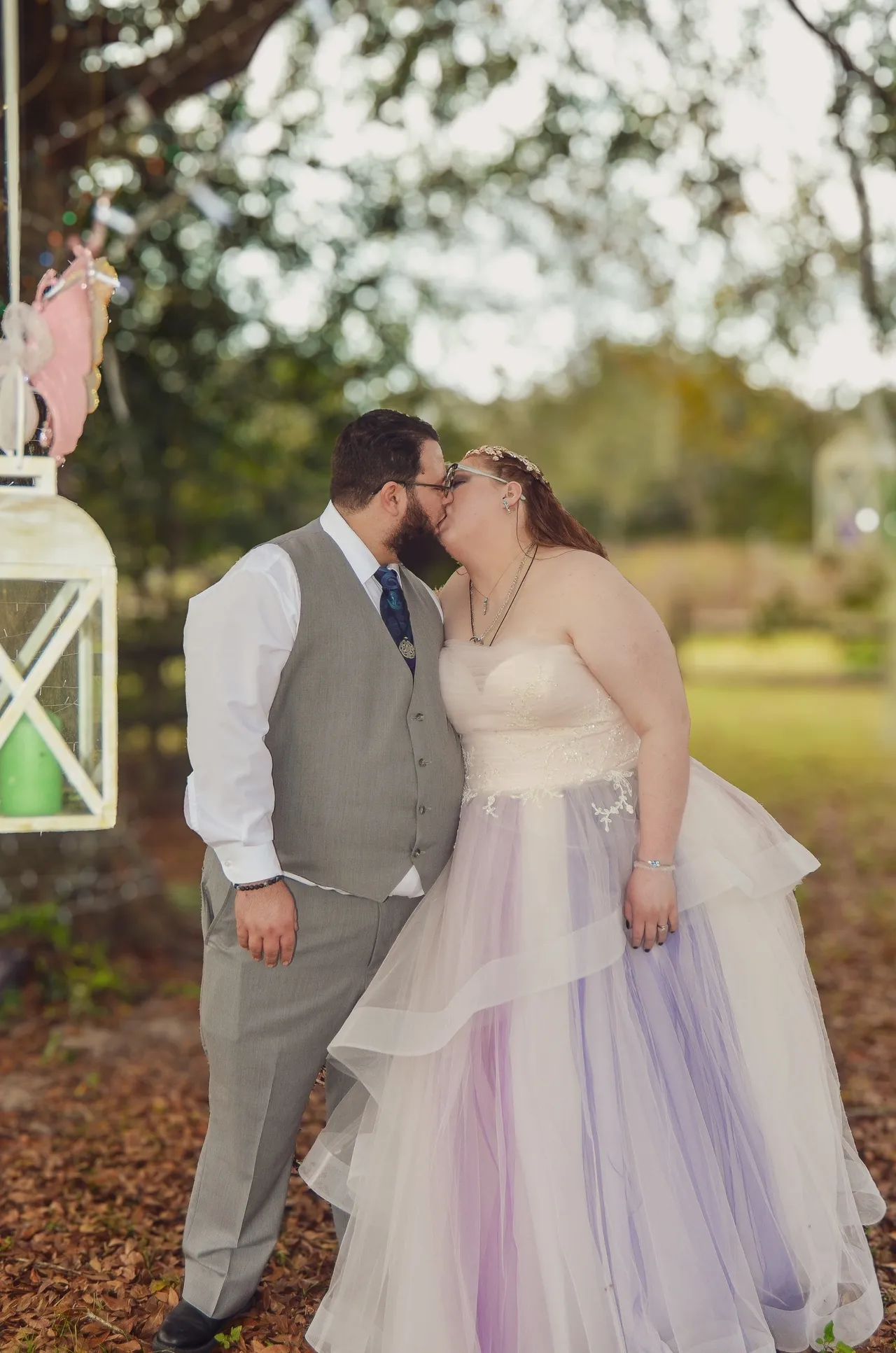 A couple kisses outdoors in a park setting; the bride wears a tiered white and lavender dress, the groom a grey vest.