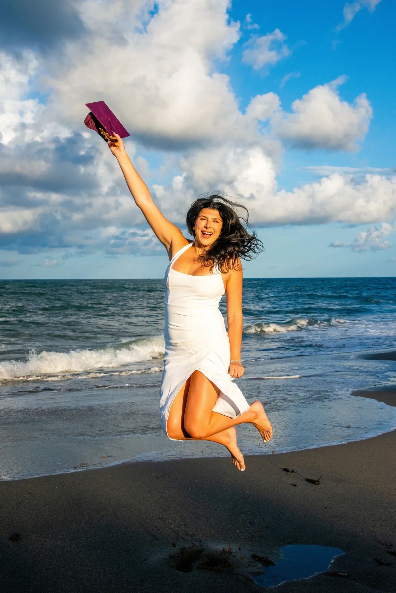 A joyful person in a white dress jumps on a beach, arm raised holding a purple object, against a cloudy blue sky.