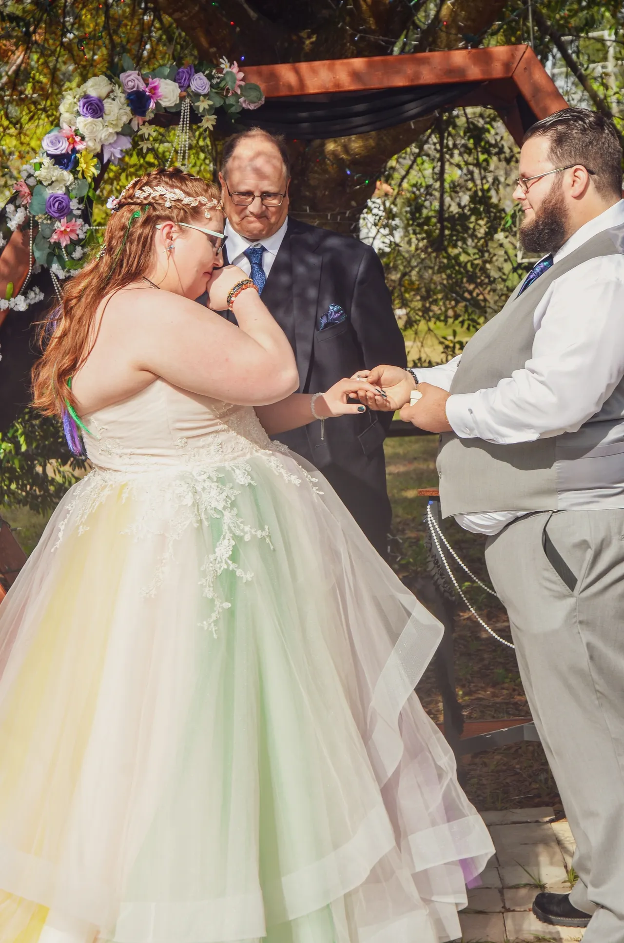 A bride in a multi-colored pastel gown cries while exchanging rings with a groom in a grey vest at an outdoor wedding.