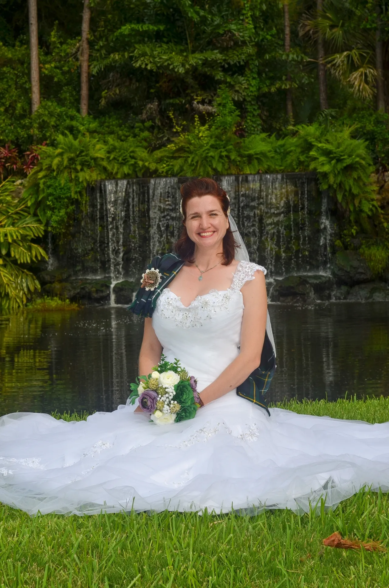 A bride in a white gown and veil holds a bouquet, sitting on grass in front of a small waterfall and pond.