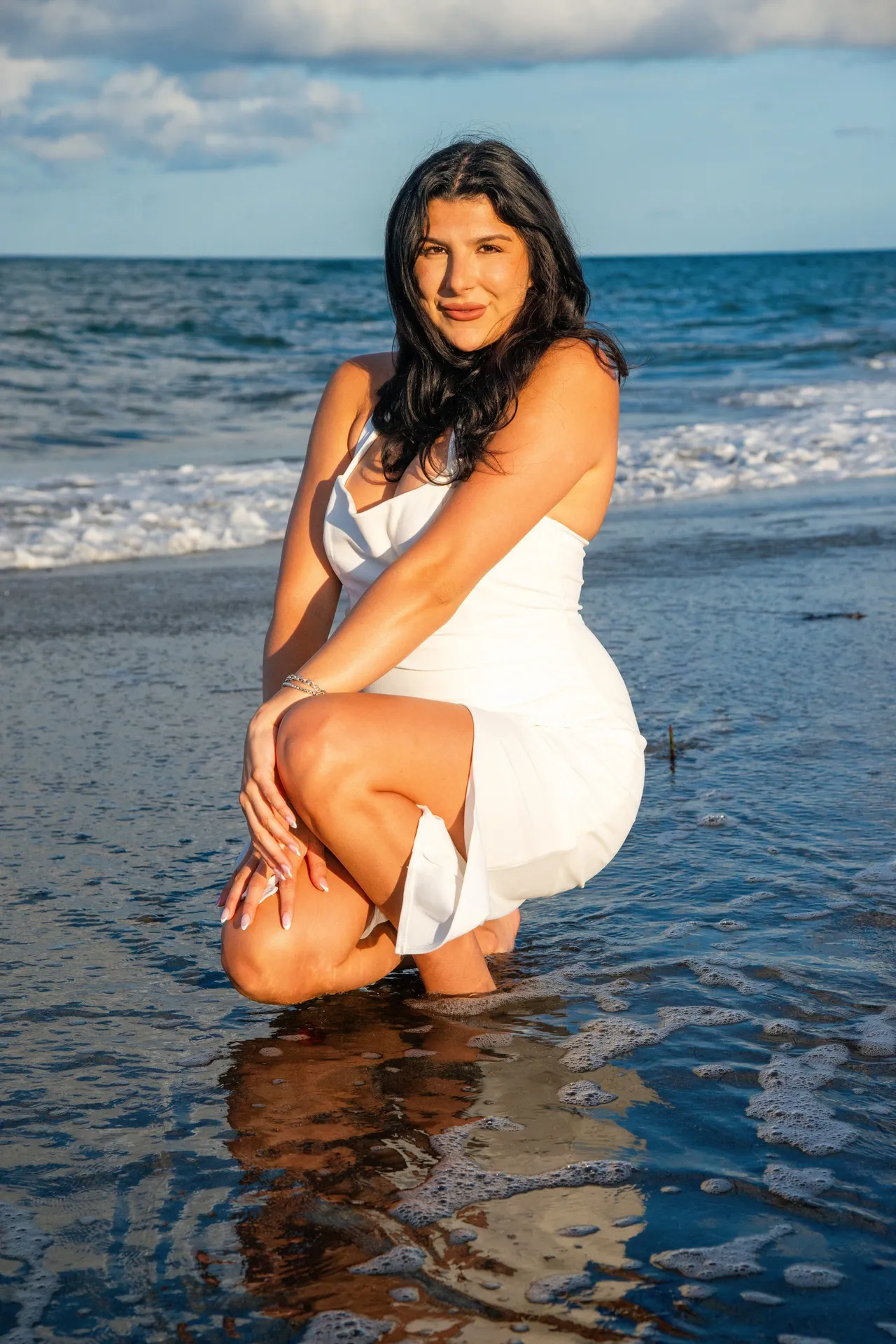 A person in a white dress crouches in shallow ocean water at the beach, smiling at the camera.