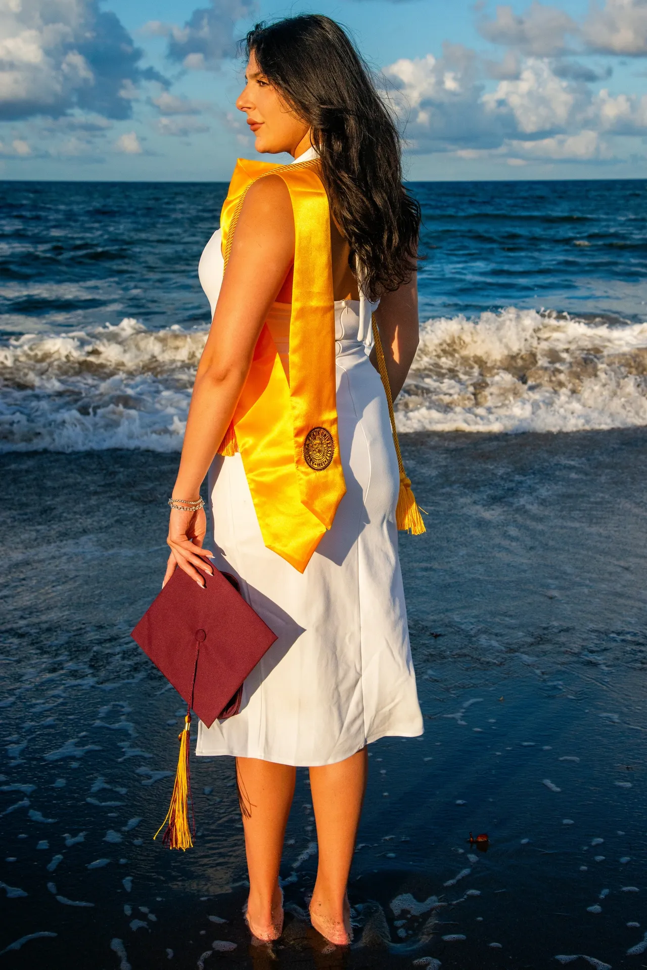 A graduate in a white dress and yellow stole stands in the ocean, holding a maroon graduation cap.