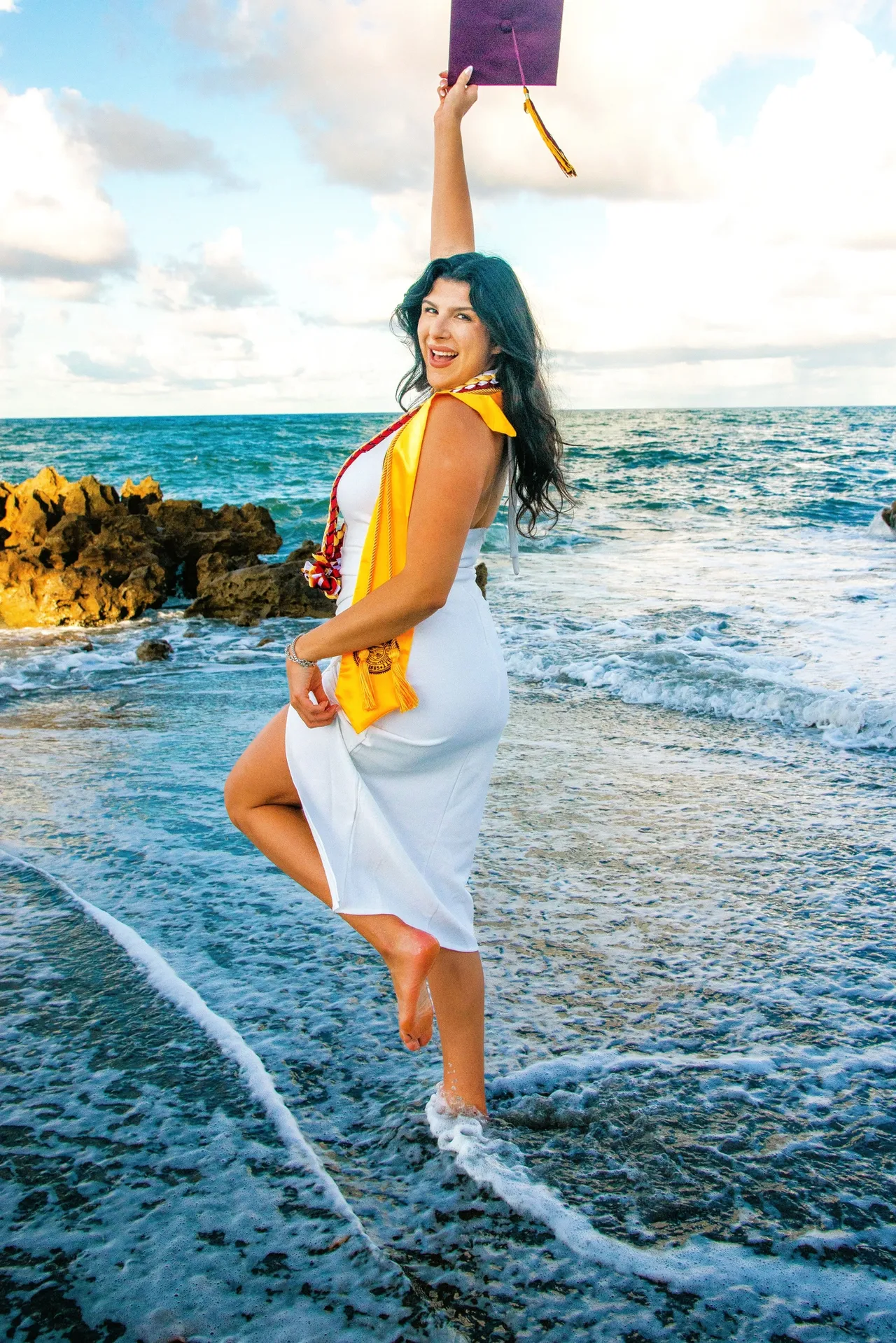 A smiling graduate in a white dress and yellow stole stands in the ocean, holding a graduation cap high in the air.