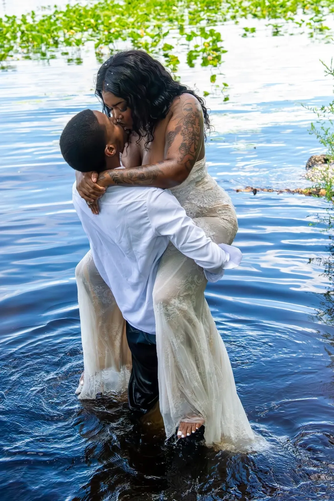 A couple embracing and kissing while standing in shallow water, the person being held wearing a long, light-colored dress.