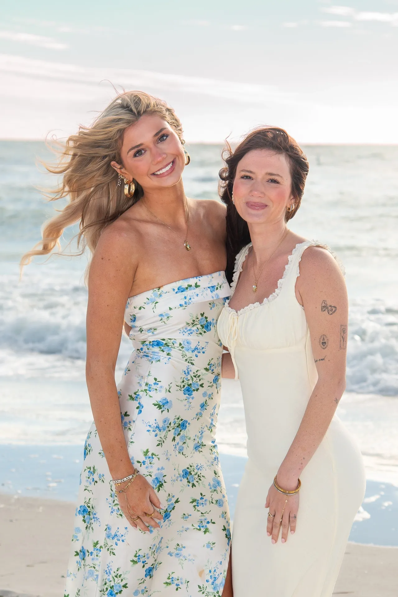 Two people in sundresses pose together on a beach at sunset, smiling toward the camera.