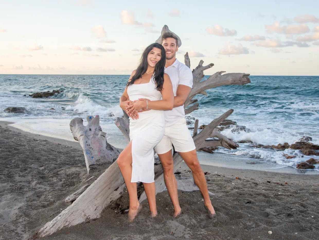 A couple in white outfits embracing in front of a large piece of driftwood on a sandy beach at sunset.