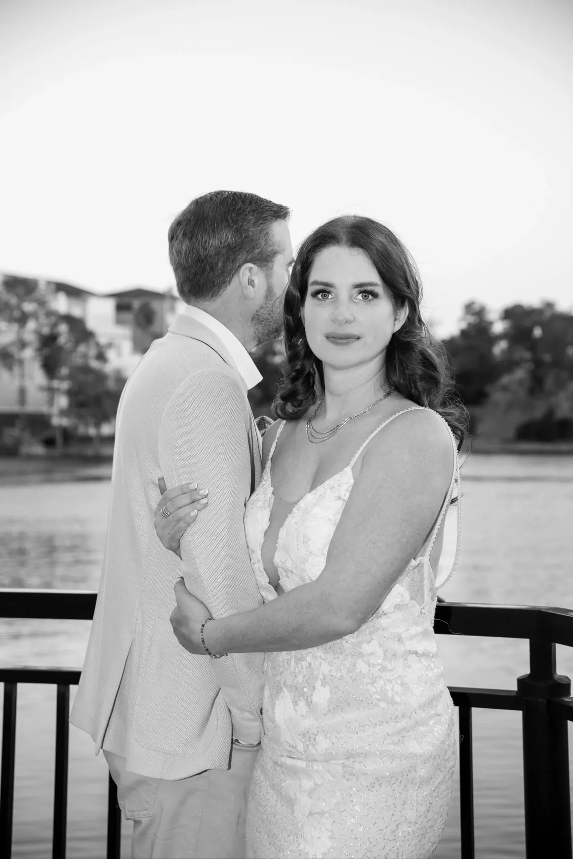 A black-and-white portrait of a couple embracing outdoors by a lake with a railing in the foreground.