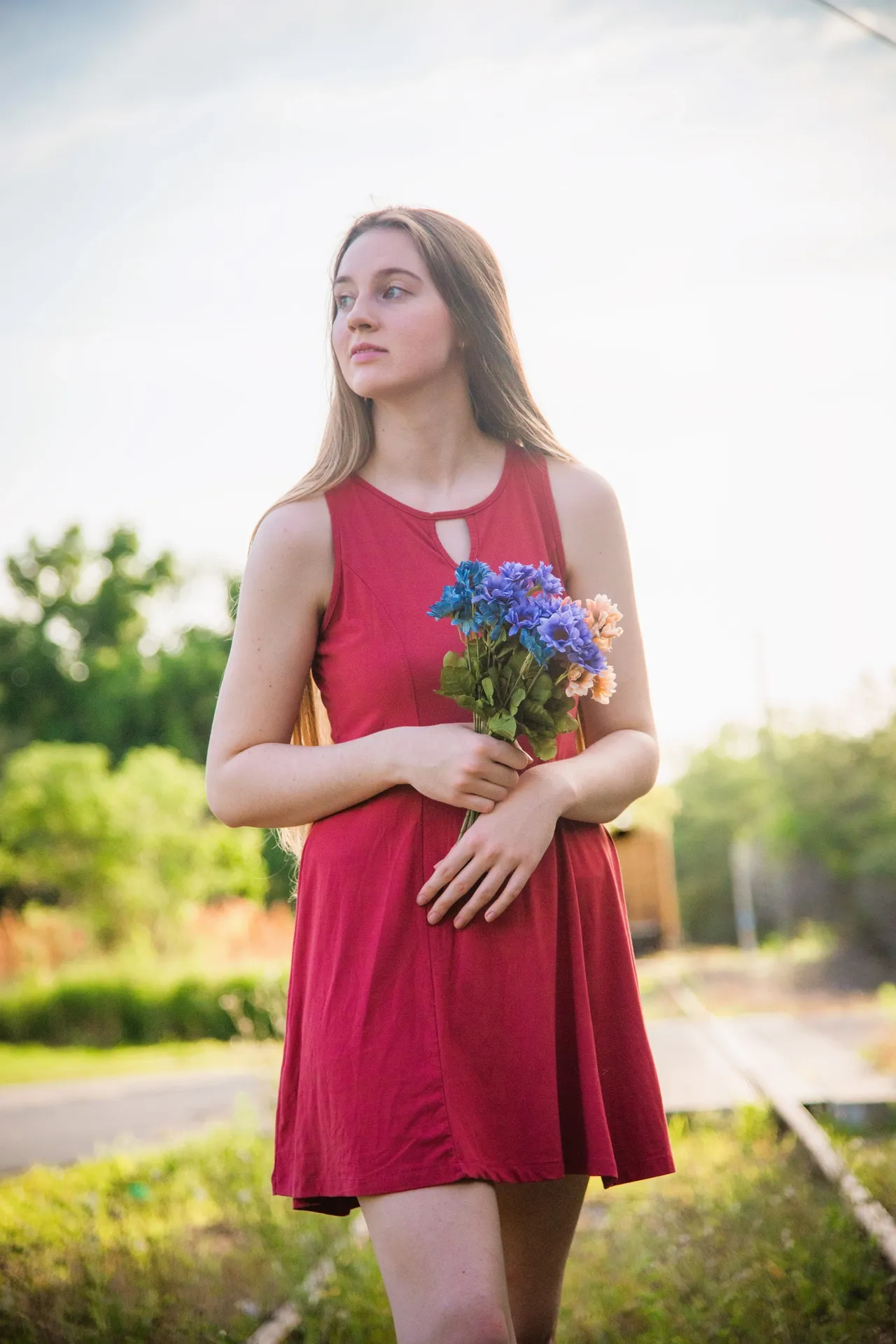 A person with long hair wearing a red dress holds a small bouquet of blue flowers while standing on outdoor tracks.