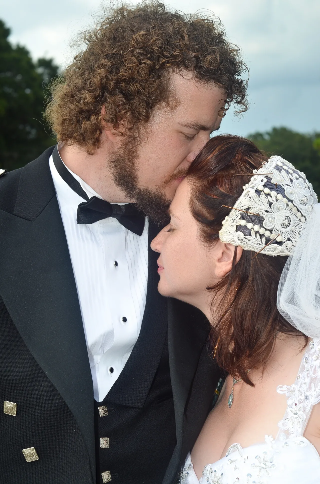 A person in a tuxedo kisses the forehead of another person in a bridal gown and a decorative lace veil.