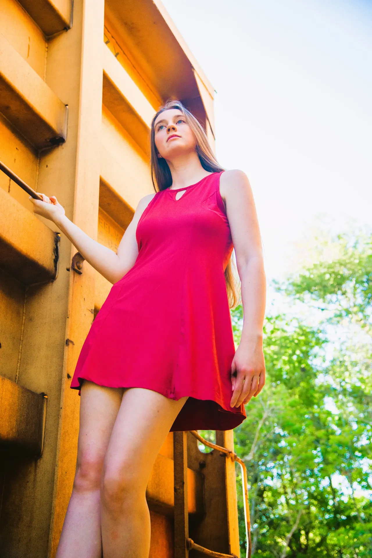 A person in a bright red mini dress stands posing beside a large, yellow industrial structure outdoors.
