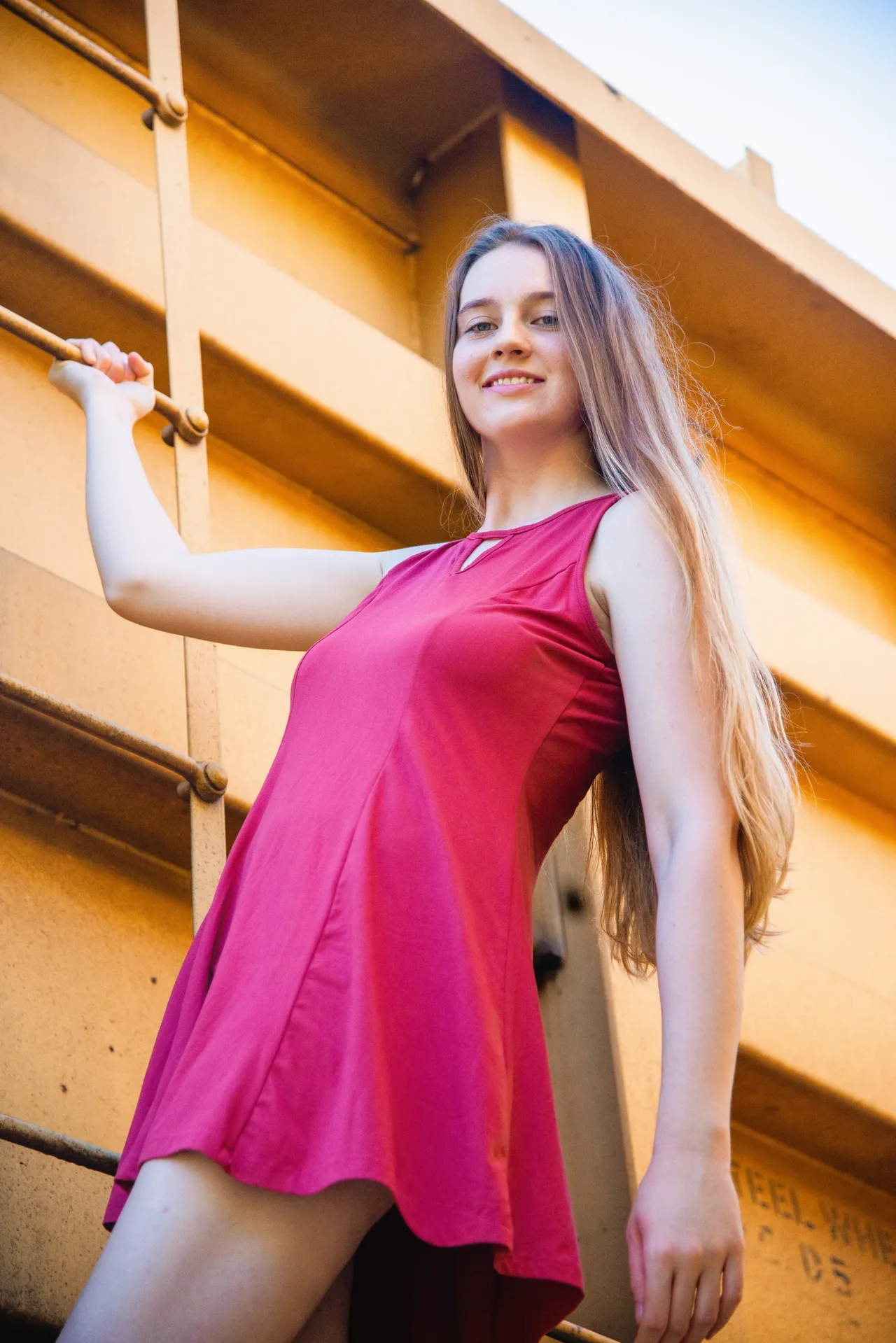 A person in a magenta dress posing against a large, yellow industrial structure with railings.