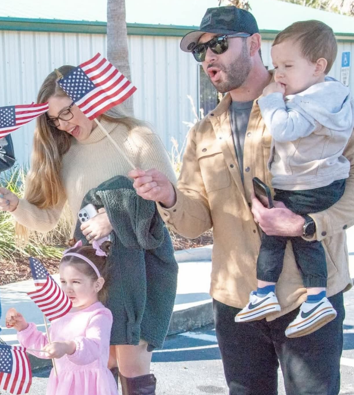 A family outdoors, holding small American flags, with the adults smiling and a child held in arms.