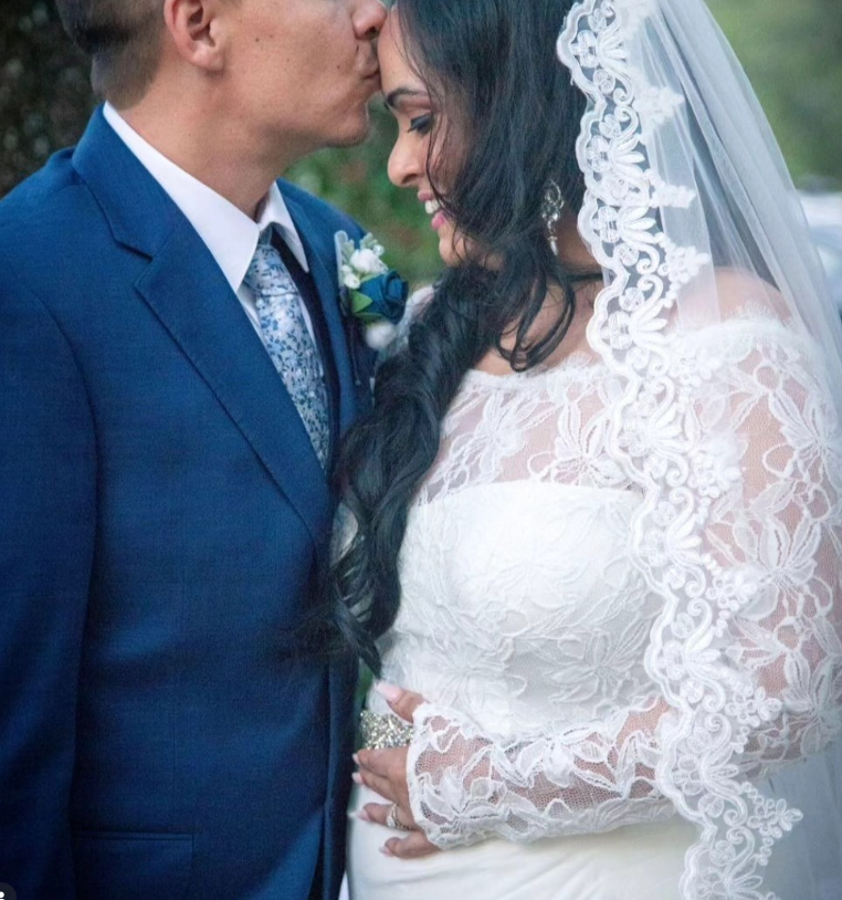 A person in a blue suit kisses the forehead of a bride in a white lace gown and veil, both smiling softly.