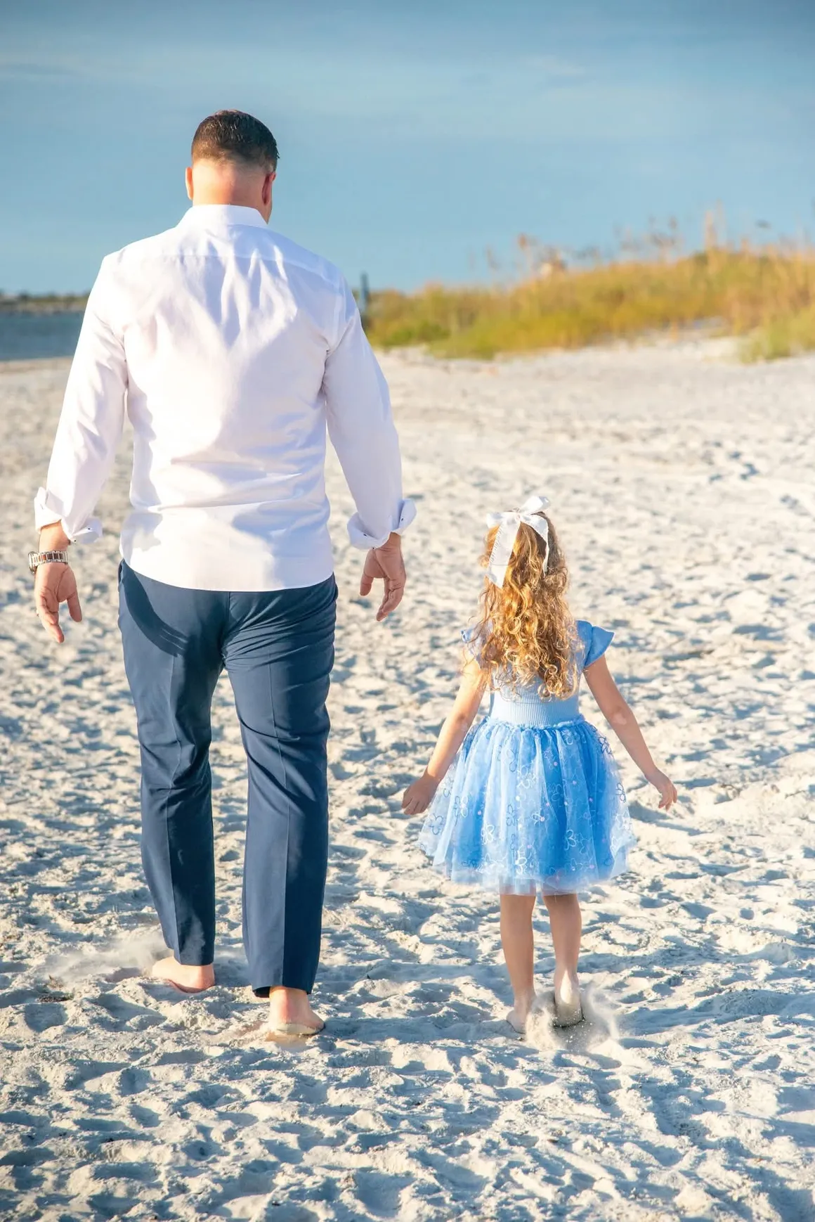 A person in a white shirt and blue trousers walks on a sandy beach next to a child in a blue tulle dress.