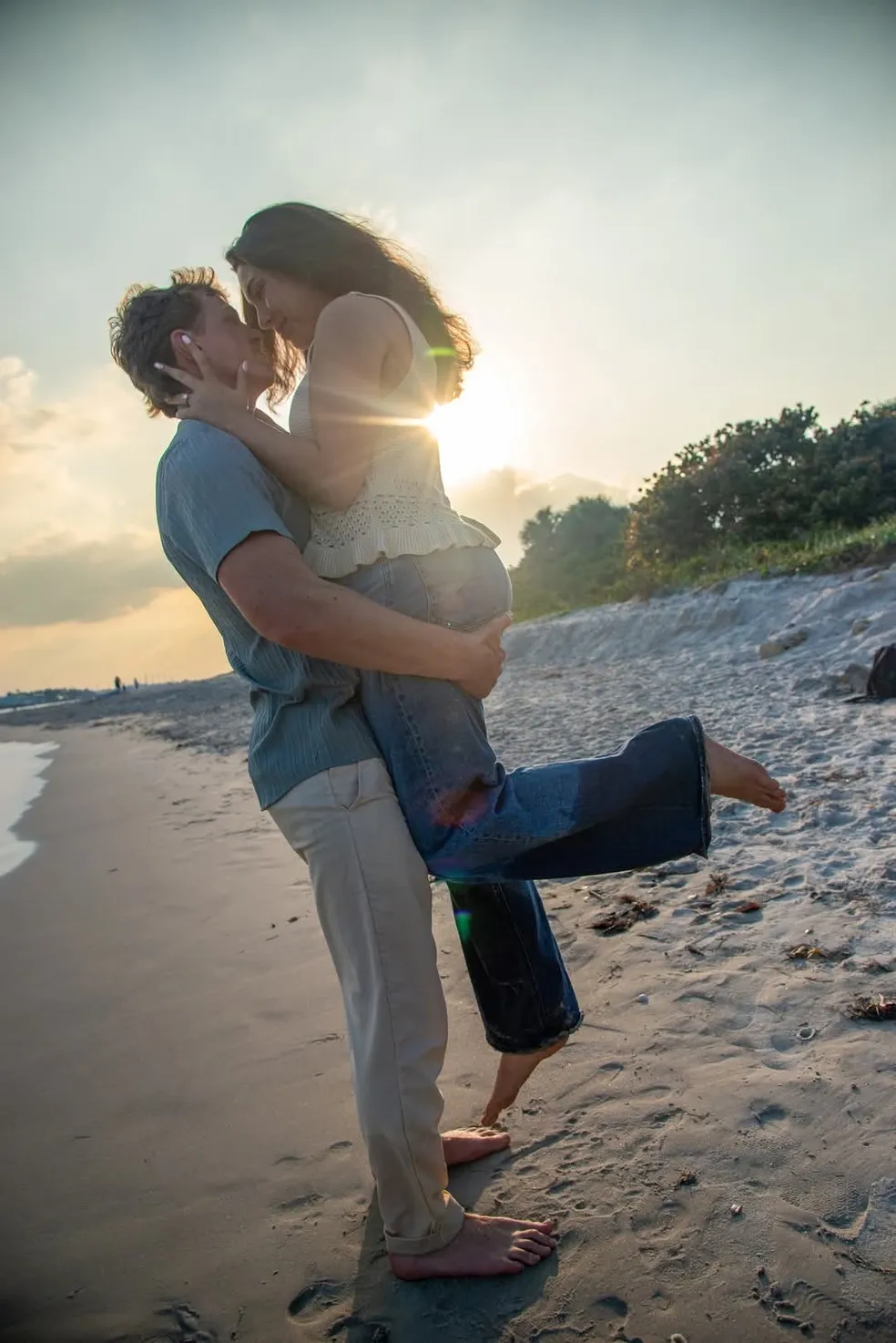 A couple embraces on a beach at sunset, with one person lifting the other in their arms.