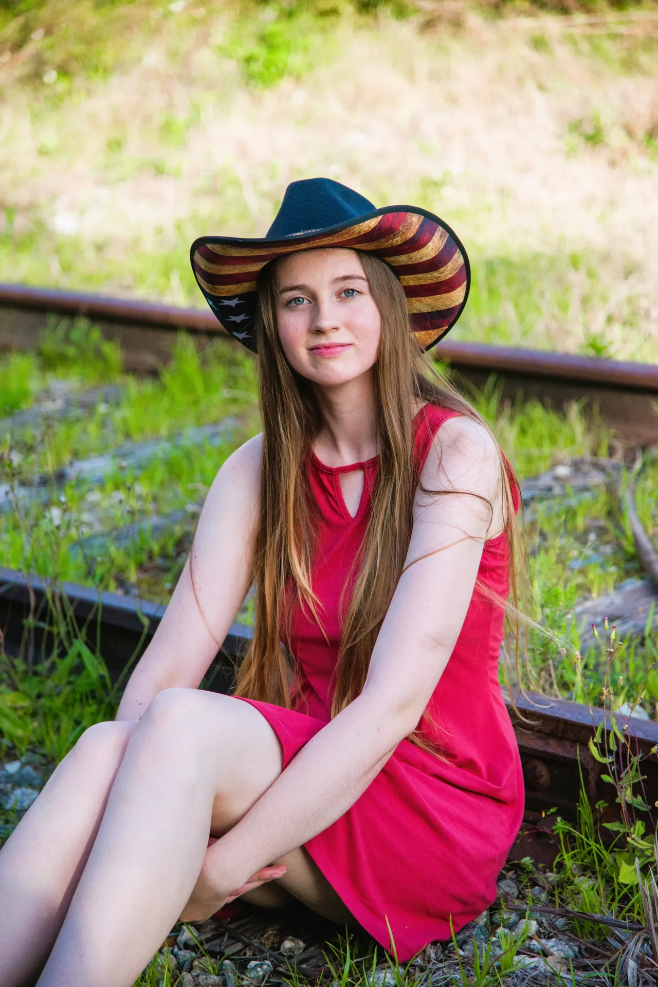 A person in a red dress and a star-and-stripes cowboy hat sits on railroad tracks surrounded by green grass.