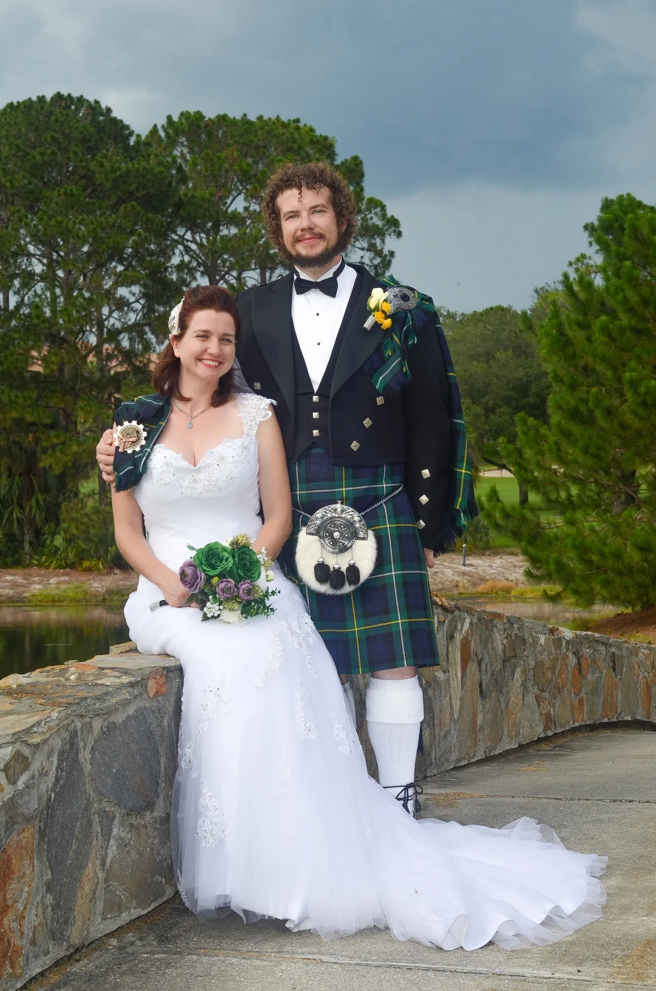 A bride in a white wedding gown and a groom in a kilt and tuxedo pose together outdoors on a stone wall.