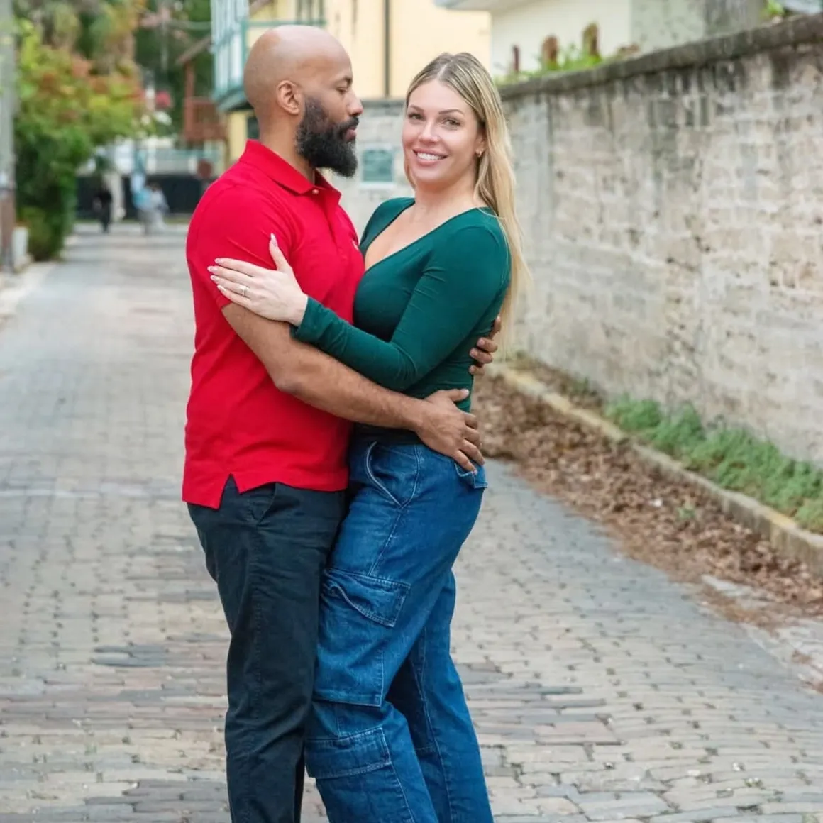 A couple embraces while standing on a cobblestone street, smiling at the camera, wearing casual clothing.