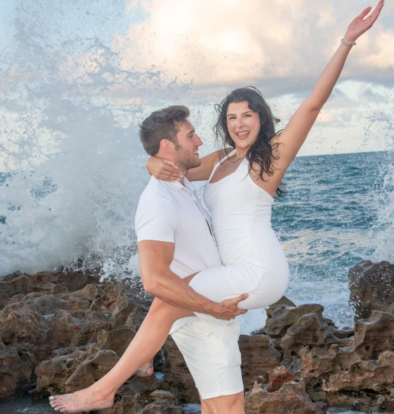 A person in white clothes holds another person in a white dress aloft on jagged rocks before a large ocean wave.