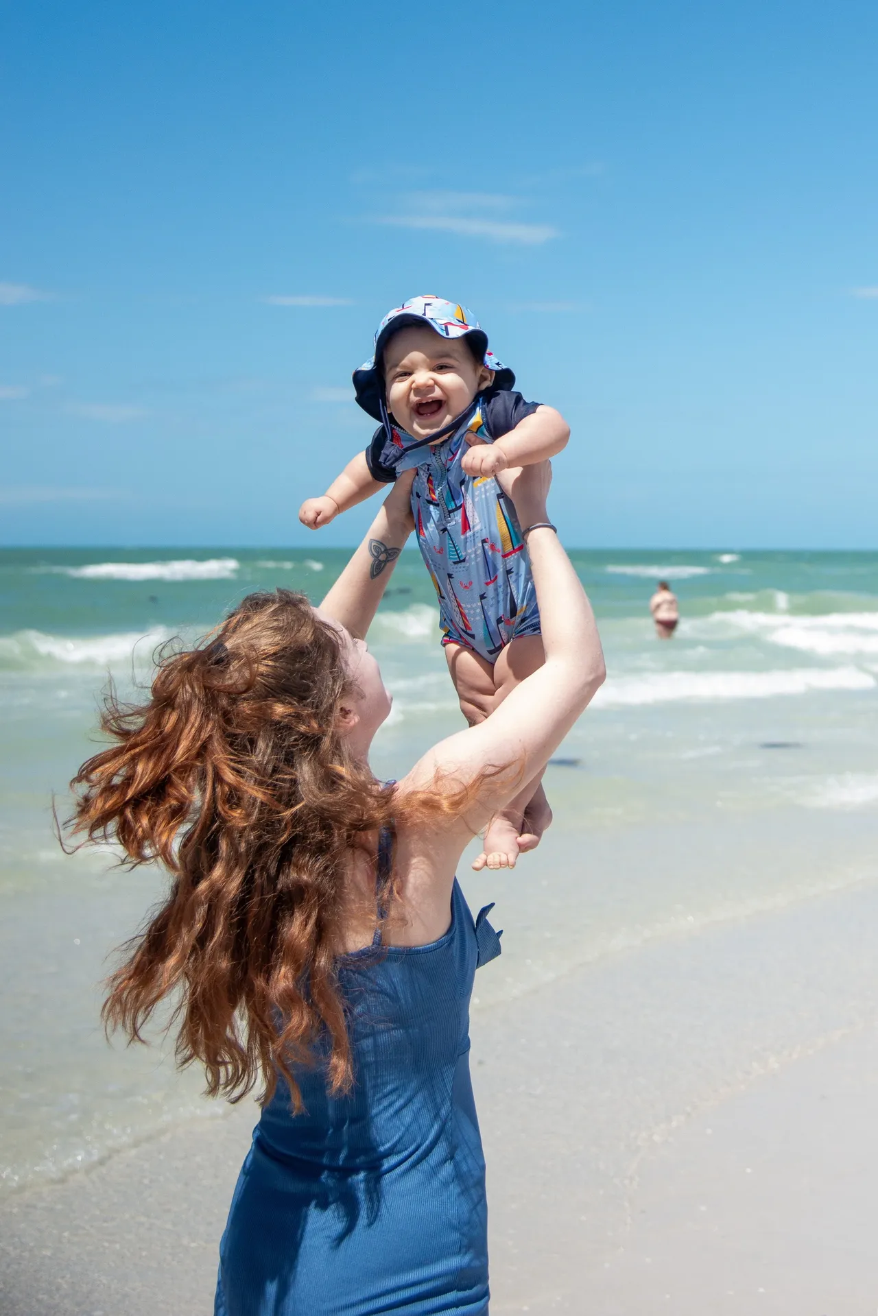 A person in a blue dress lifts a happy infant in a matching sun hat and romper high above them on a sunny beach.