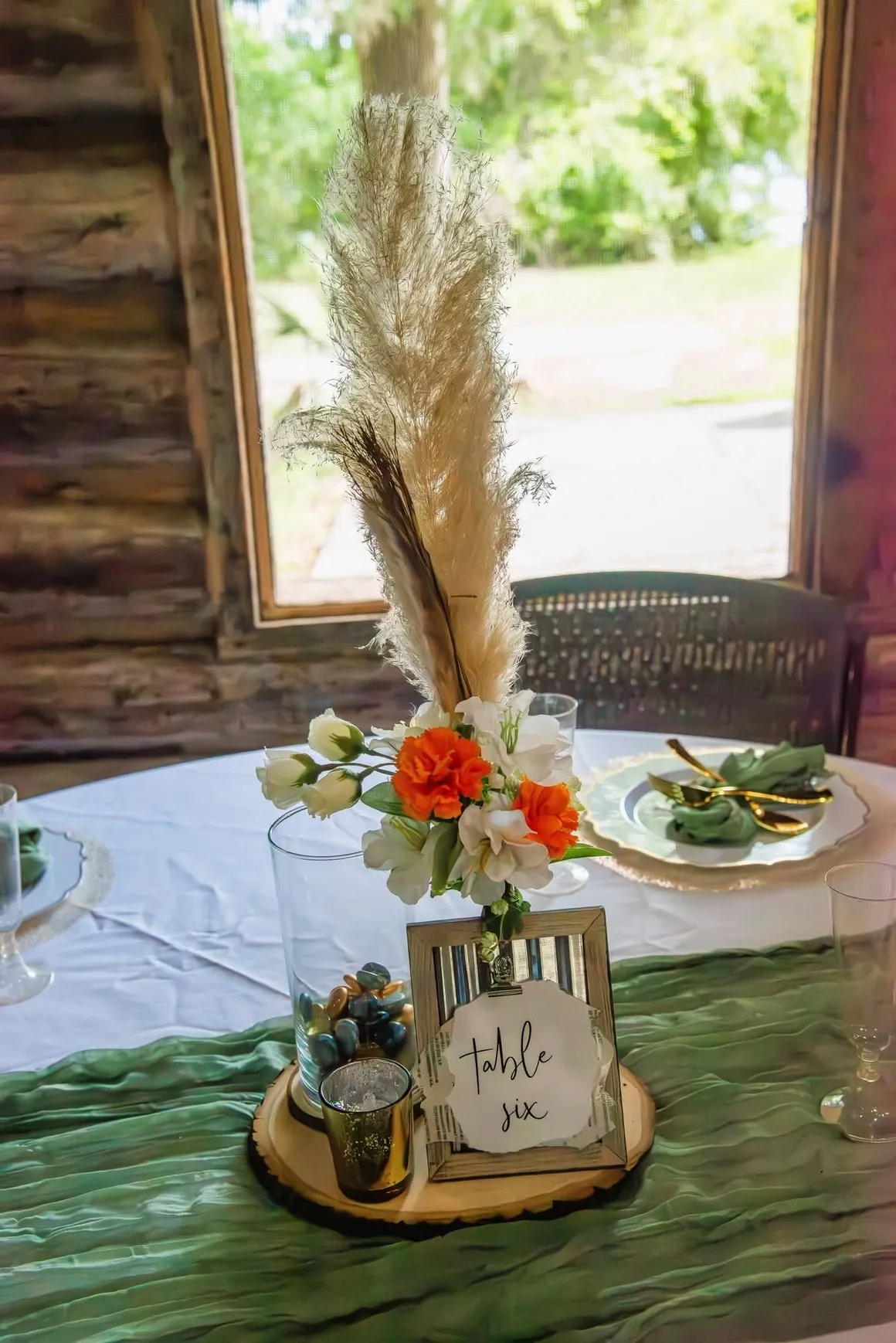 A table centerpiece with pampas grass, white and orange flowers, and a 'Table Six' sign on a wood slice runner.