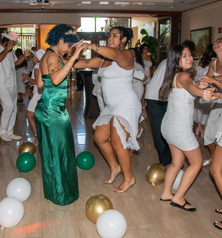 People dance together at an indoor party, wearing formal white and green attire among scattered balloons on the floor.