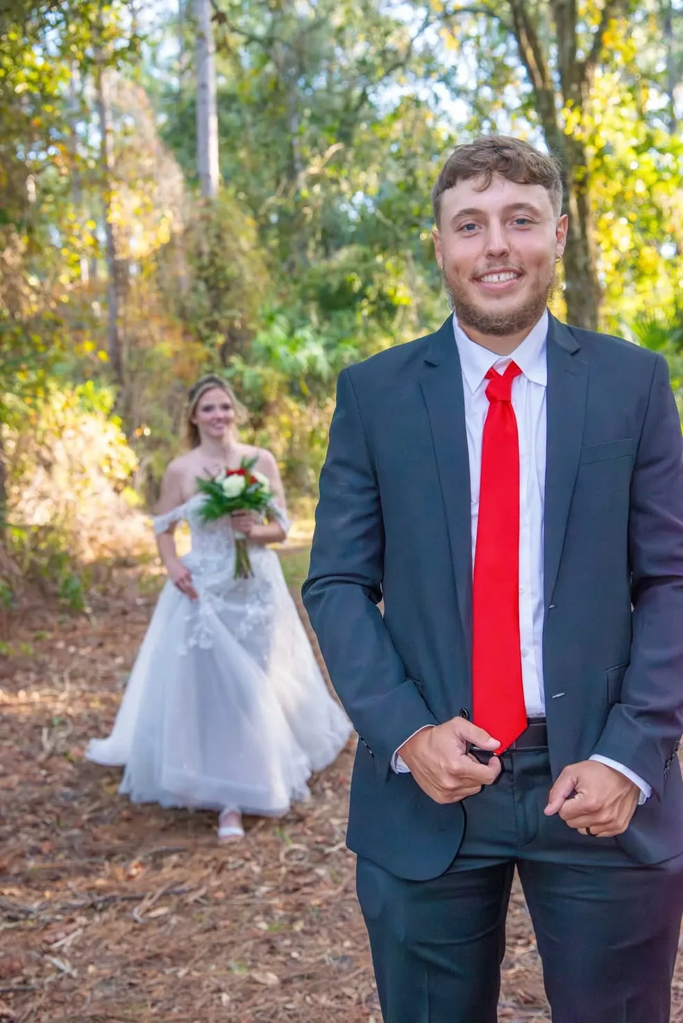 A person in a dark suit with a red tie smiles in the foreground while a person in a wedding dress approaches in a forest.