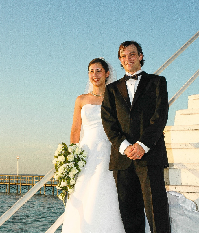 A bride and groom stand smiling together on white stairs overlooking a pier and the ocean at sunset.