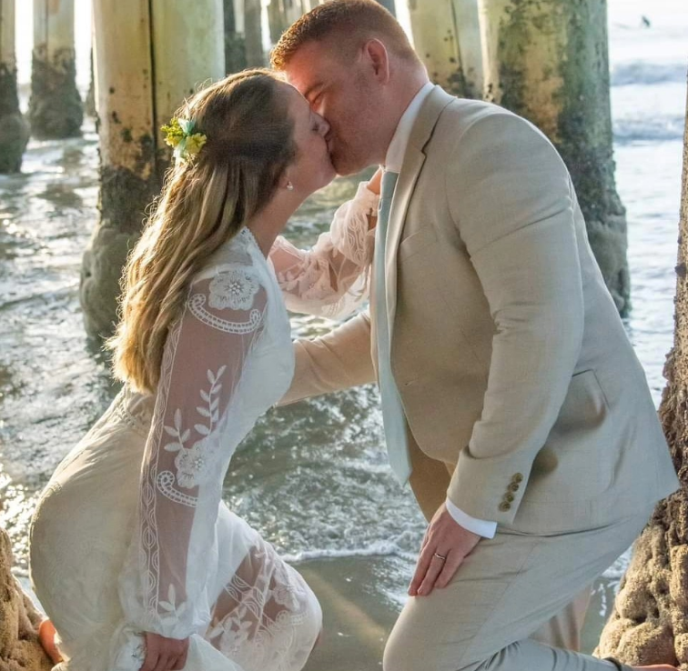 A couple in wedding attire kisses while standing in the shallow water under a pier.