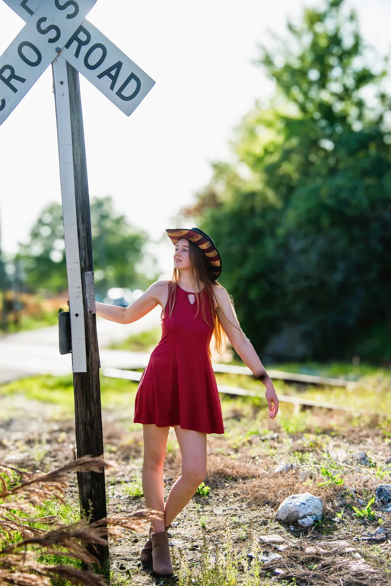 A person in a red dress and hat leaning against a railroad crossing sign in a grassy, outdoor setting.