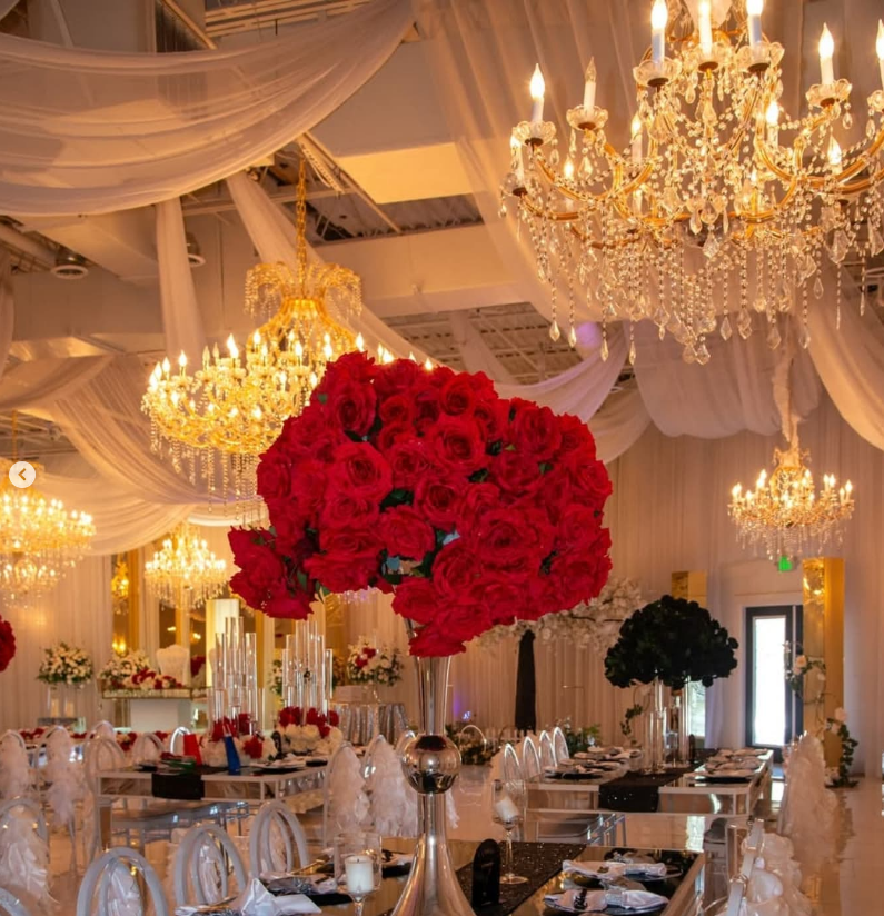 A formal event hall decorated with draped white fabric, crystal chandeliers, and large centerpieces of red roses.