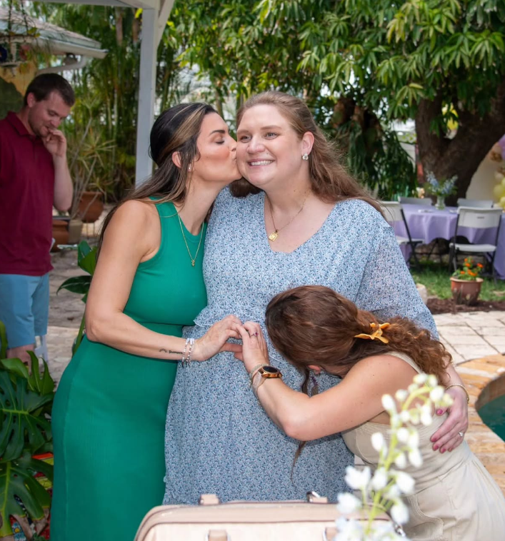 A woman in a green dress kisses a smiling, pregnant woman, while another person leans in to embrace her stomach.