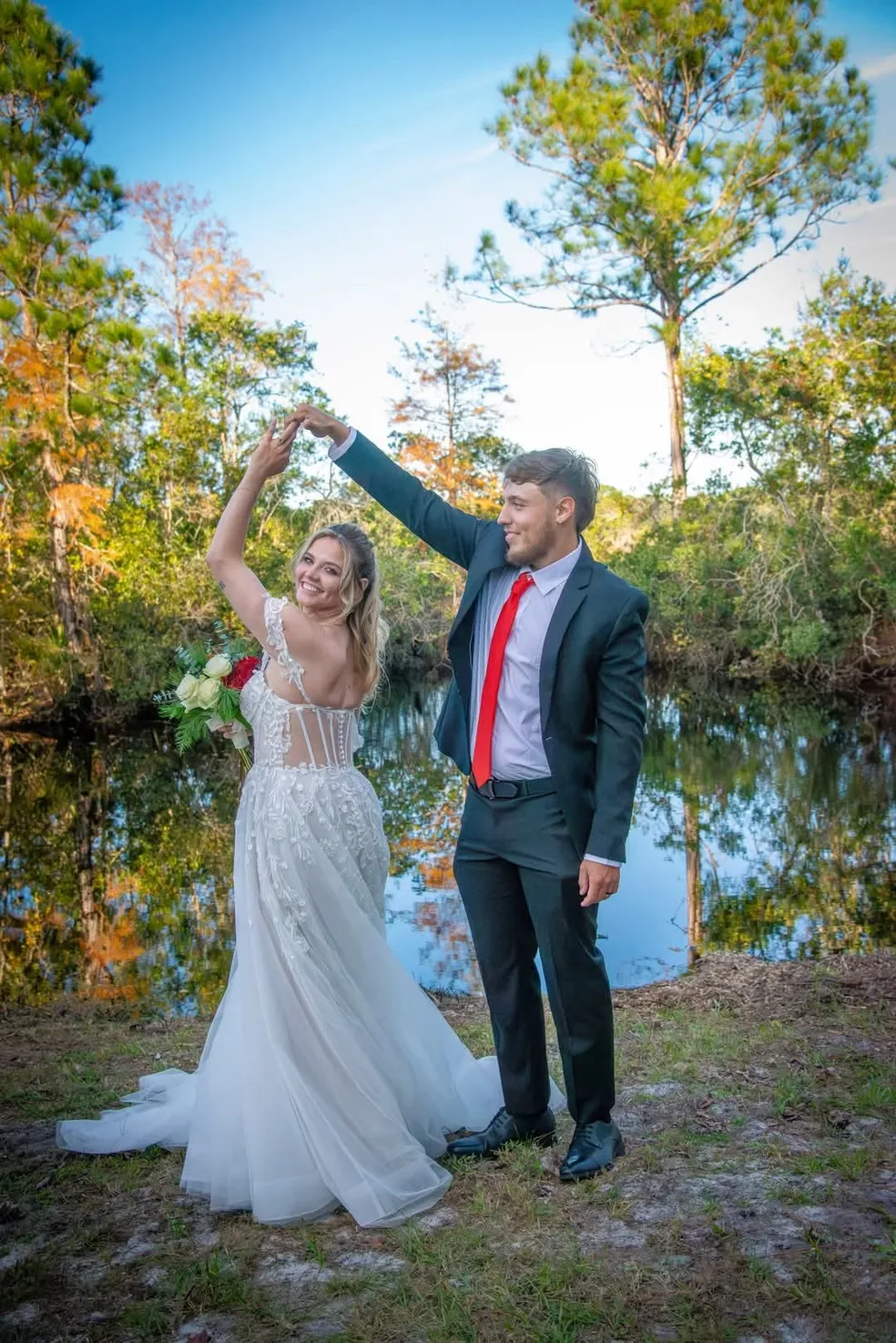 A bride in a white wedding gown and a groom in a dark suit with a red tie pose while dancing by a pond in a wooded area.