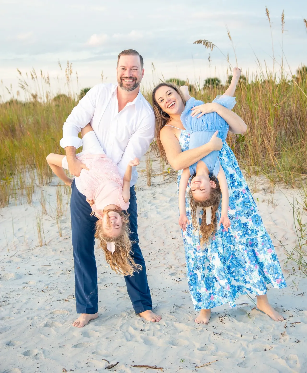 Parents hold two children upside down while standing on a sandy beach during a sunny day.